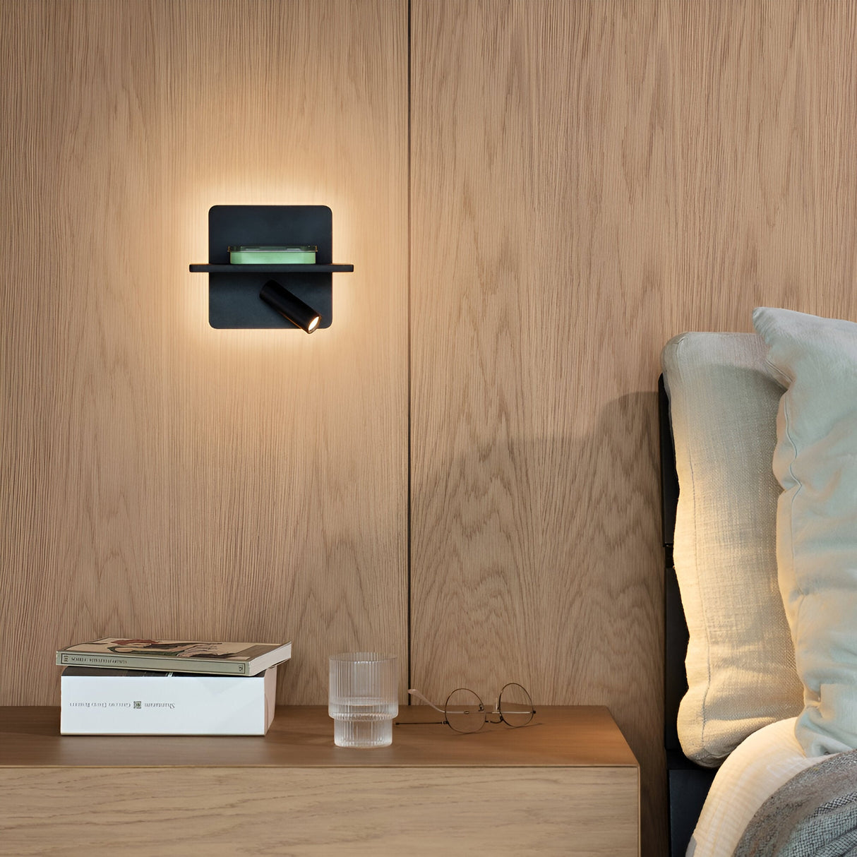 A contemporary bedroom display features a wooden wall and headboard, highlighted by the Gleon LED Wall Light 11.5W 3000K - Black, which tastefully lights a small shelf holding a book. Below, an integrated USB port is available on the wooden nightstand with another book, glasses, and a textured glass. A bed with white pillows is partially visible in the scene.