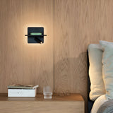 A contemporary bedroom display features a wooden wall and headboard, highlighted by the Gleon LED Wall Light 11.5W 3000K - Black, which tastefully lights a small shelf holding a book. Below, an integrated USB port is available on the wooden nightstand with another book, glasses, and a textured glass. A bed with white pillows is partially visible in the scene.