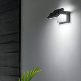 The Solstice LED Outdoor Wall Light 20.5W 3000K - Anthracite casts a luminous, angular glow on the white surface. Its anthracite finish creates a sophisticated contrast with dark wood paneling, while a small green plant in the corner introduces a touch of nature to the contemporary design.