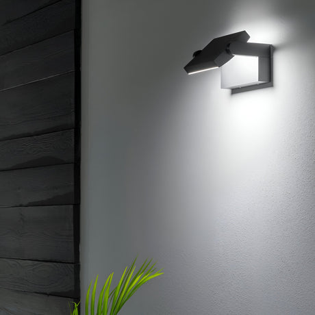 The Solstice LED Outdoor Wall Light 20.5W 3000K - Anthracite casts a luminous, angular glow on the white surface. Its anthracite finish creates a sophisticated contrast with dark wood paneling, while a small green plant in the corner introduces a touch of nature to the contemporary design.