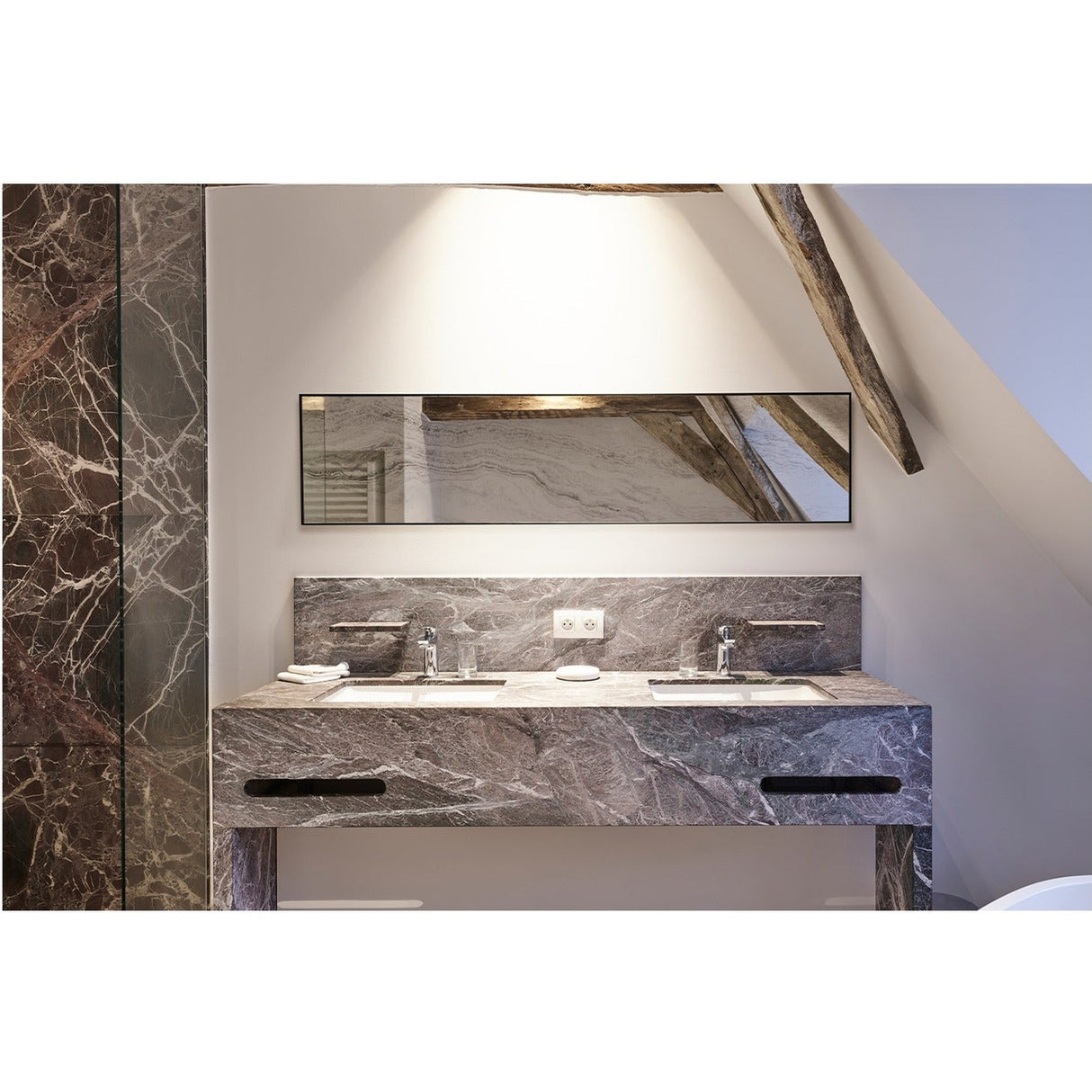 Travertin rectangular wall mirror with thin black metal frame, mounted above a marble double-sink vanity in a bathroom with sloped wooden beams.