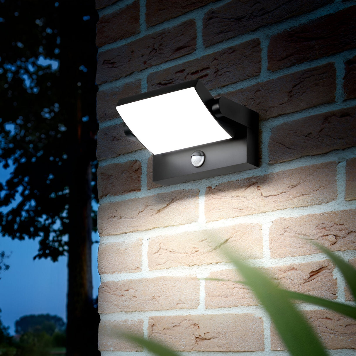 The Solstice LED Outdoor Motion Sensor Wall Light 21W 3000K in anthracite is mounted on a brick wall. It emits a warm glow against the dark background, with blurred green leaves adding a serene touch to the foreground.