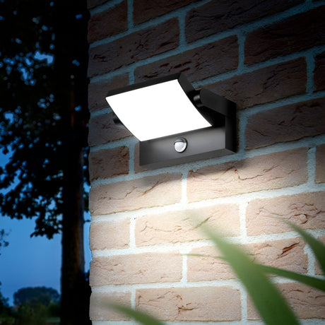 The Solstice LED Outdoor Motion Sensor Wall Light 21W 3000K in anthracite is mounted on a brick wall. It emits a warm glow against the dark background, with blurred green leaves adding a serene touch to the foreground.