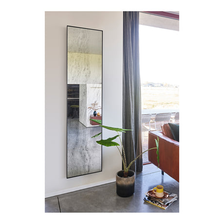 Travertin tall rectangular wall mirror with thin black metal frame and travertine-print glass, shown on a living room wall beside a curtained window with a potted plant nearby.