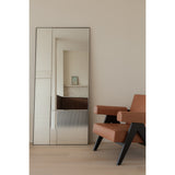 Finestra Flutes XL full-length mirror in a black metal frame, leaning against a wall in a living room, combining fluted textured glass with a plain mirror panel beside a tan armchair.