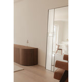 Finestra Flutes full-length mirror in a black metal frame, split between fluted textured glass and plain mirror panels, leaning in a living room beside a walnut sideboard.