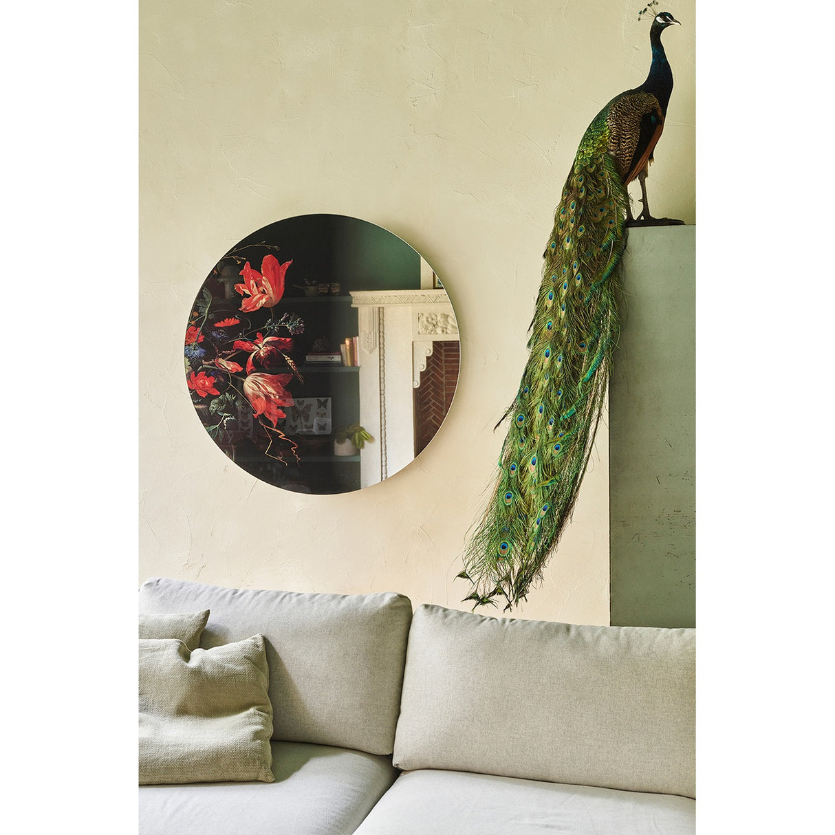 Museo Still Life Printed Round Wall Mirror 100cm, frameless glass with dark floral still-life print, hung above a beige sofa beside a taxidermy peacock display.