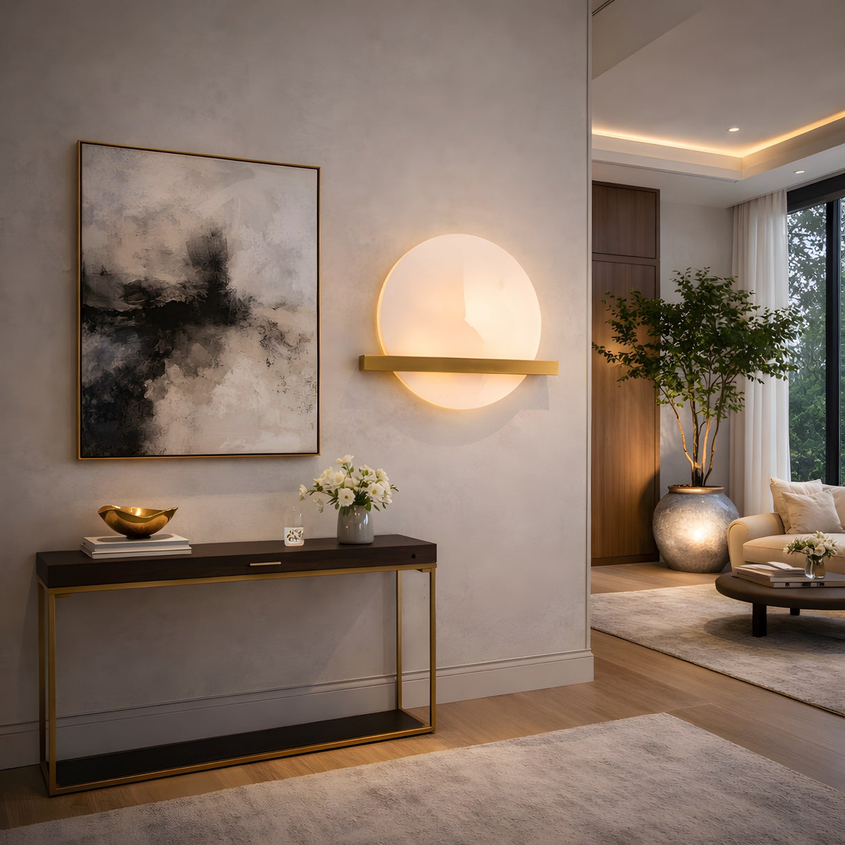A modern living room with minimalist decor, showcasing a console table with flowers, a gold bowl, abstract wall art, the Caldris LED Circular Alabaster Wall Light in Brushed Brass & Soft White, a potted tree, and neutral furnishings in bright light.