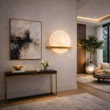 A modern living room with minimalist decor, showcasing a console table with flowers, a gold bowl, abstract wall art, the Caldris LED Circular Alabaster Wall Light in Brushed Brass & Soft White, a potted tree, and neutral furnishings in bright light.