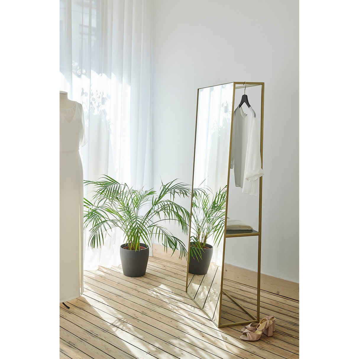 Chassis XL Deluxe free standing cheval mirror in a gold open metal frame with a marble shelf, styled in a bedroom holding a hanger with clothing beside a plant and sheer curtains.