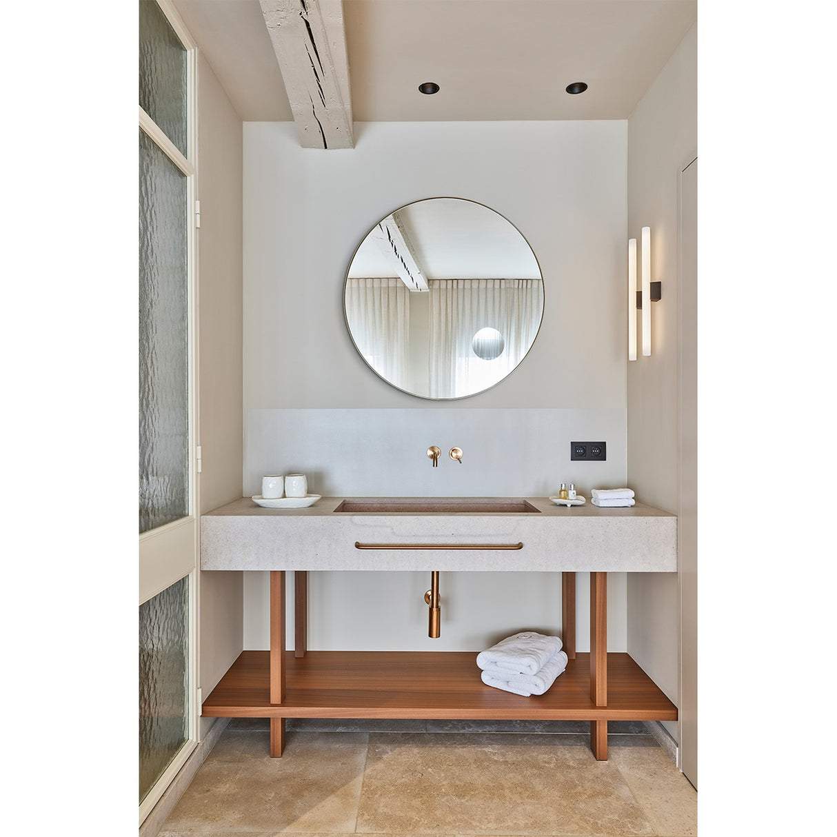 Round bronze-framed wall mirror with built-in magnifying section above a bathroom vanity with wood-legged stone counter, brass taps, and a round mirror reflecting curtained window.