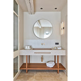 Round bronze-framed wall mirror with built-in magnifying section above a bathroom vanity with wood-legged stone counter, brass taps, and a round mirror reflecting curtained window.