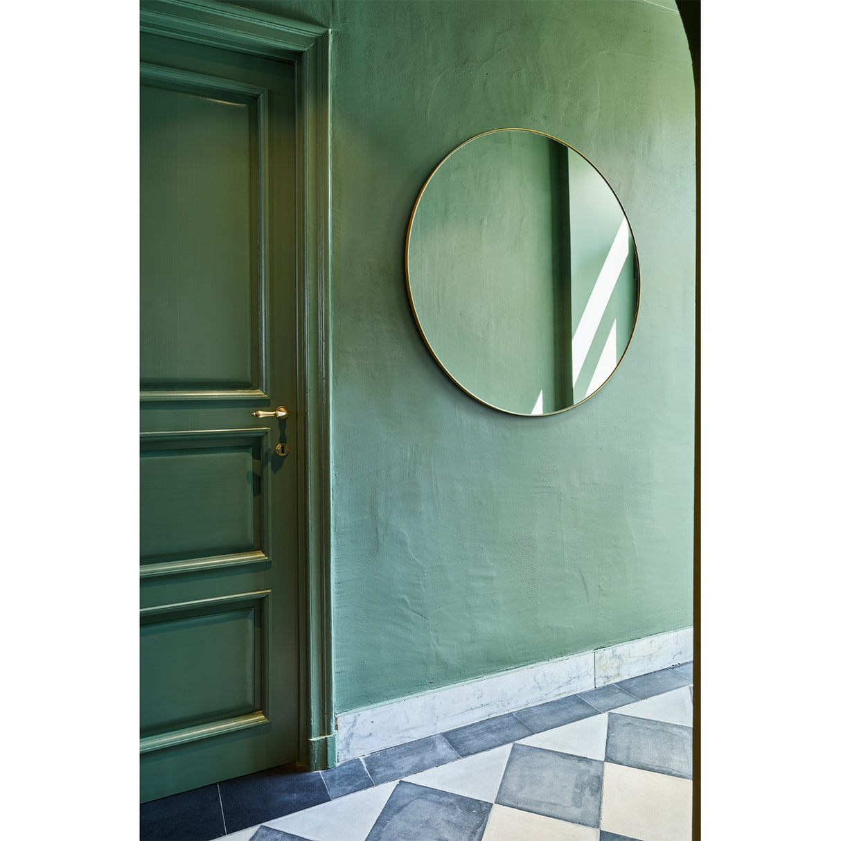 Hoop Bronze Round Wall Mirror 105cm with a thin bronze metal frame, hung on a green wall beside a green panelled door in a hallway with patterned floor tiles.
