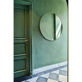 Hoop Bronze Round Wall Mirror 105cm with a thin bronze metal frame, hung on a green wall beside a green panelled door in a hallway with patterned floor tiles.