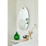 Globo Lago Oak Peg Hung Wall Mirror, a frameless organic-shaped mirror with a bevelled edge, hung from a solid oak peg on a light bedroom wall above a shelf with decor.