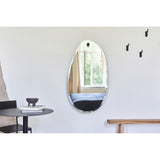 Frameless organic-shaped Globo Lago wall mirror with bevelled edge, hung from a solid oak peg on a light grey wall in a living room setting.