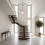 A minimalist modern entryway features a floating wooden staircase, glass railing, marble accent wall, console table with decor, and floor-to-ceiling windows—all illuminated by the Esmira 1 Light Layered Dome Alabaster Pendant in soft white & brushed brass.