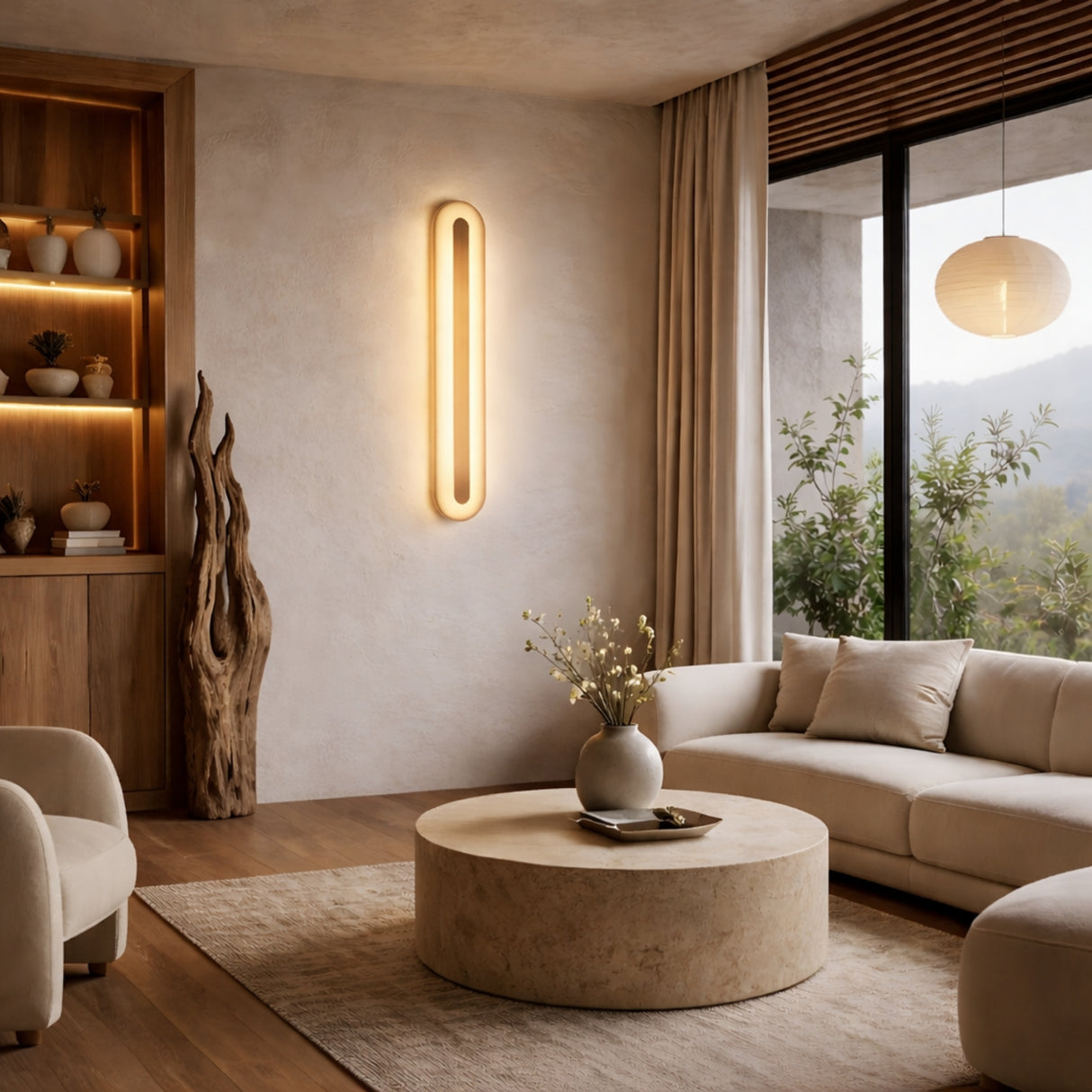 A modern living room with neutral tones features a round beige coffee table, light sofas, sculptural floor lamp, built-in shelving, and the Averra LED Large Round Alabaster Wall Light in Soft White & Brass illuminating the space.