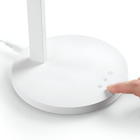 A hand grazes the circular button on the base of the Arcanta LED Table Lamp - White. This modern lighting piece features touch-sensitive controls with icons for power and light adjustment. Its cord extends from the back, providing functionality alongside an adjustable diffuser for a customized ambiance.