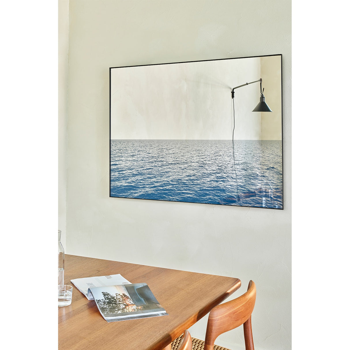 Maro Seascape Printed Glass Wall Mirror 120cm in a thin black frame, hung above a wooden dining table, with a printed blue seascape below and mirrored glass reflecting a wall lamp above.
