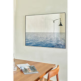 Maro Seascape Printed Glass Wall Mirror 120cm in a thin black frame, hung above a wooden dining table, with a printed blue seascape below and mirrored glass reflecting a wall lamp above.