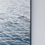 Maro Seascape Printed Glass Wall Mirror 120cm, close-up of the rectangular black-framed edge showing printed blue seascape against a plain light grey wall.