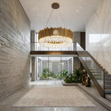 A modern, spacious foyer with concrete and marble walls, a stunning Royal 6-12 Crystal Light Chandelier - Gold, glass stairs with metal railing, indoor plants, a beige rug, and abundant natural light from large windows and a central courtyard.