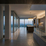 Modern open-plan kitchen with floor-to-ceiling windows and wood cabinetry, highlighted by a striking Magari 9 Light Chandelier - Gold above the large island.