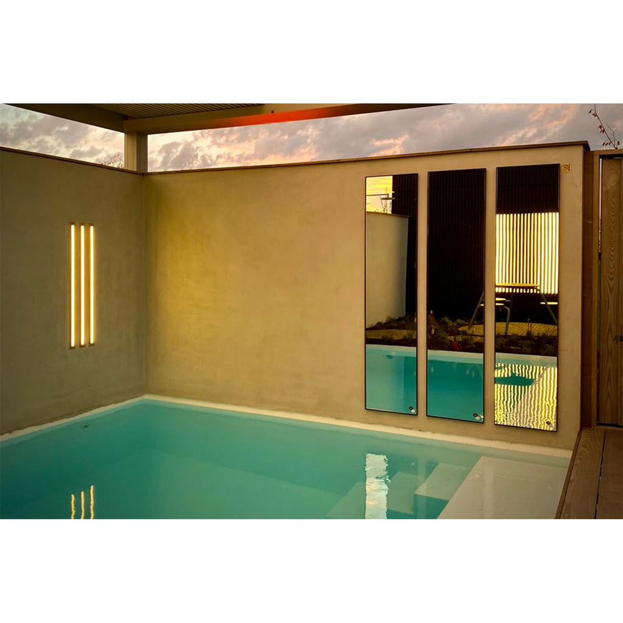 Three Lucka Outdoor full-length rectangular mirrors with slim black metal frames mounted on a poolside wall, reflecting the sunset sky and outdoor seating area.