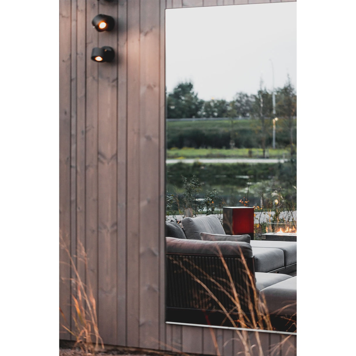 Lucka Outdoor Full Length Garden Mirror 175cm in black, slim metal frame mounted on wood cladding, reflecting an outdoor lounge area with a lake and greenery beyond.