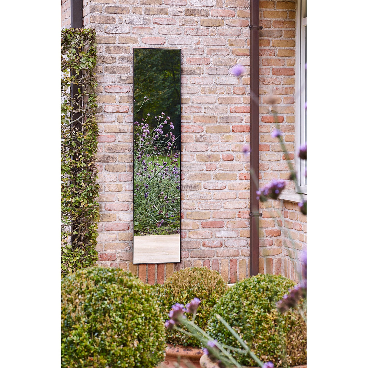 Lucka Outdoor Full Length Garden Mirror 175cm in black, slim metal frame mounted on a brick wall, reflecting greenery and purple flowers in a garden setting.