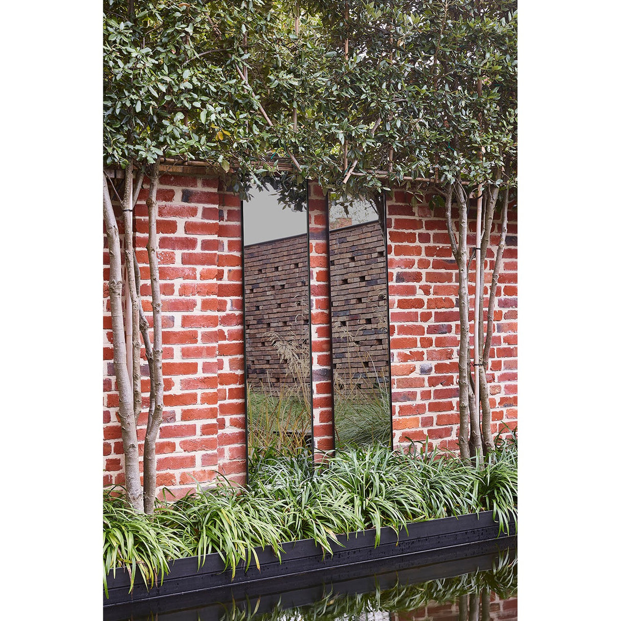 Lucka Outdoor Full Length Garden Mirror 175cm in black, slim metal frame mounted on a red brick garden wall amid trees, grasses and a reflecting pool.