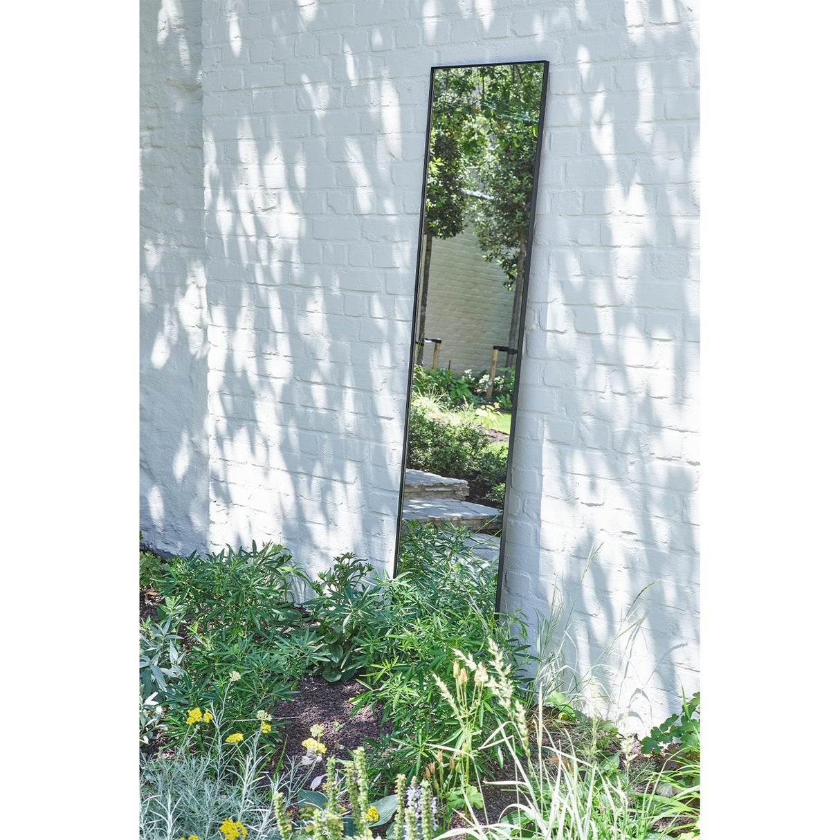 Lucka Outdoor Full Length Garden Mirror 175cm in black, slim metal frame, leaning against a white brick wall in a garden setting with greenery below.