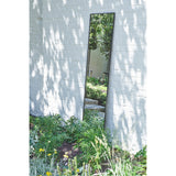 Lucka Outdoor Full Length Garden Mirror 175cm in black, slim metal frame, leaning against a white brick wall in a garden setting with greenery below.