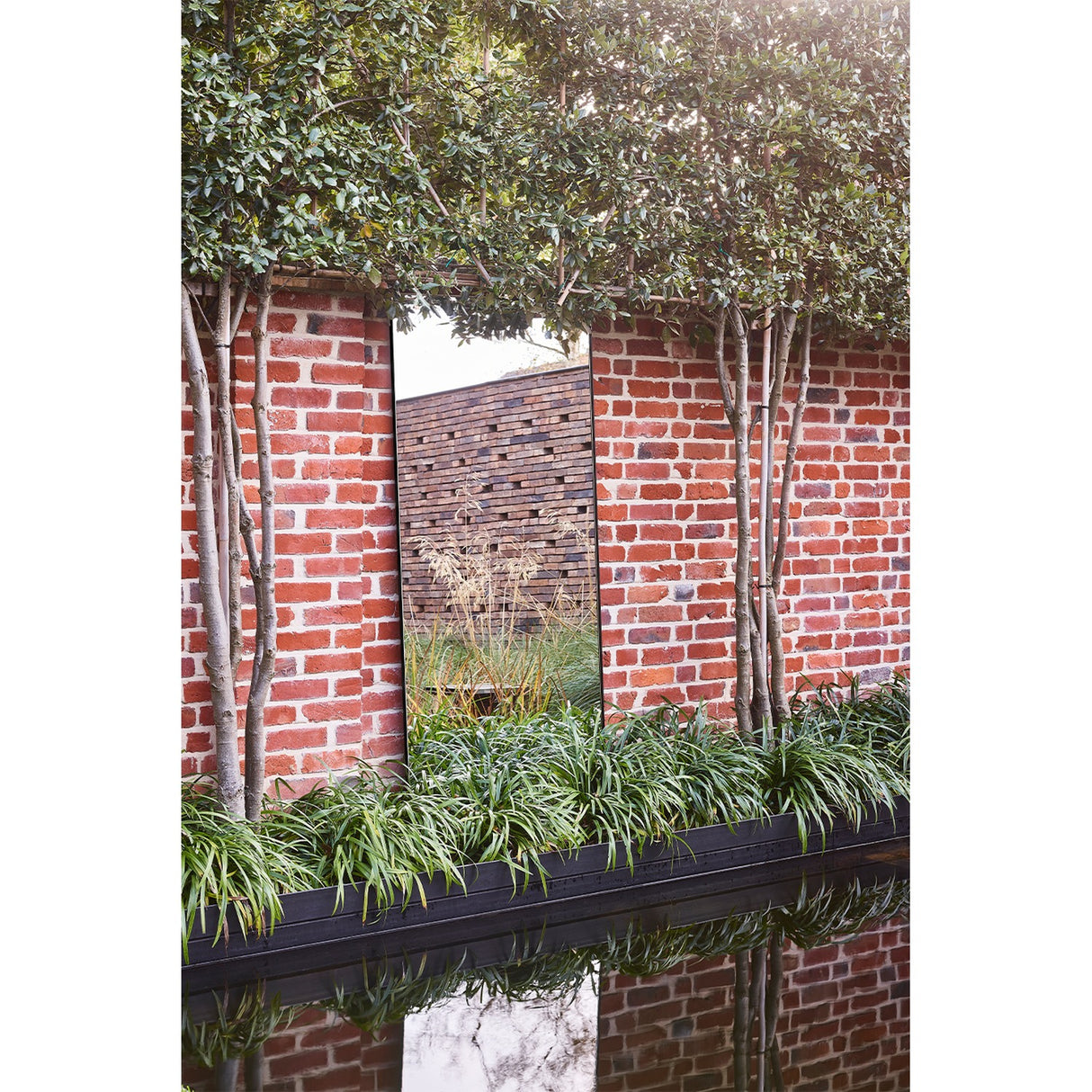 Lucka Outdoor XL full length garden mirror, 175cm, slim black metal frame, mounted on a red brick wall reflecting trees and greenery.
