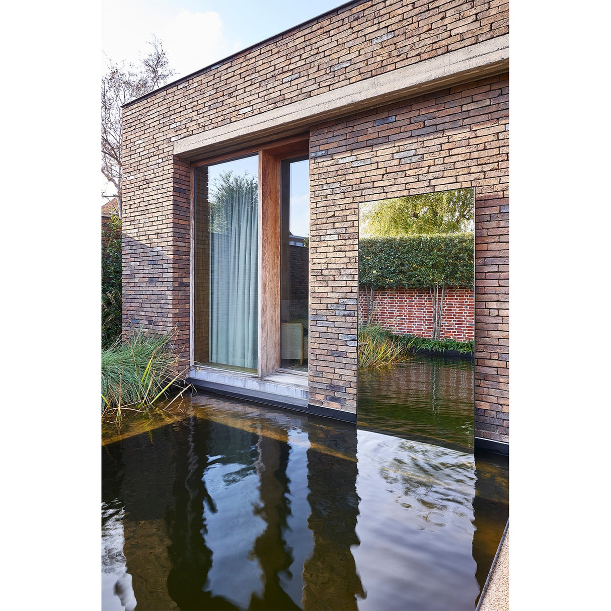 Lucka Outdoor XL full length garden mirror in black metal frame, mounted on a brick wall beside a glazed door, reflecting hedges above a still water feature.