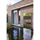Lucka Outdoor XL full length garden mirror in black metal frame, mounted on a brick wall beside a glazed door, reflecting hedges above a still water feature.
