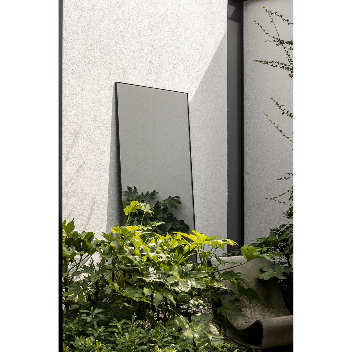 Lucka Outdoor XL full length garden mirror in a slim black metal frame, leaning against a light grey exterior wall beside lush green foliage.