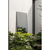 Lucka Outdoor XL full length garden mirror in a slim black metal frame, leaning against a light grey exterior wall beside lush green foliage.