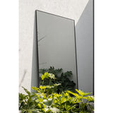 Lucka Outdoor XL Full Length Garden Mirror 175cm in a slim black metal frame, leaning against a grey outdoor wall beside green foliage.