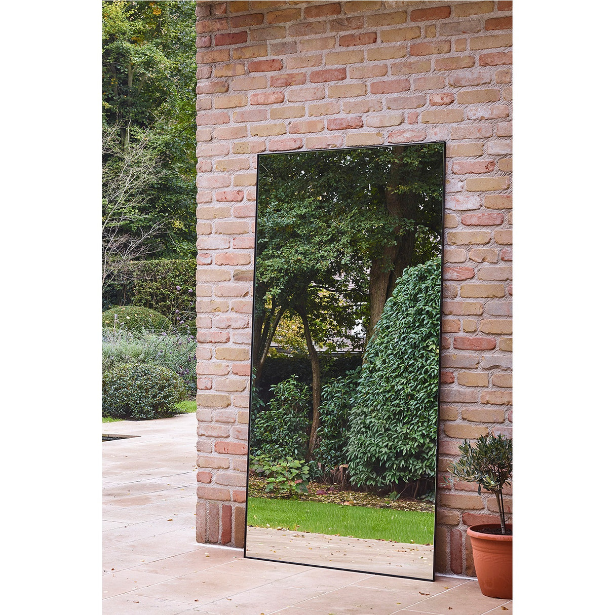 Lucka Outdoor XL full length mirror, 175cm, slim black metal frame, leaning against a brick wall outdoors and reflecting trees and greenery in a garden setting.