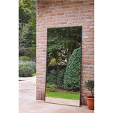 Lucka Outdoor XL full length mirror, 175cm, slim black metal frame, leaning against a brick wall outdoors and reflecting trees and greenery in a garden setting.