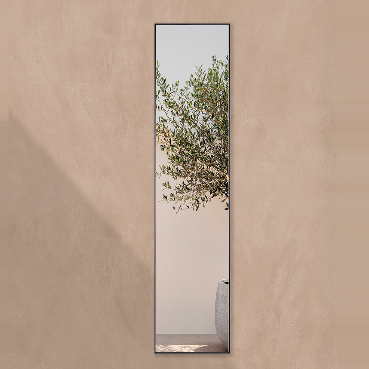 Slim full length outdoor mirror in a satin black frame on a terracotta plaster garden wall, reflecting an olive tree.