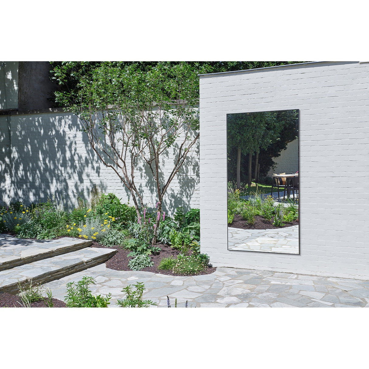 Lucka Outdoor XXL full length garden mirror, 200cm rectangular black metal frame, mounted on a white brick exterior wall reflecting garden greenery and a stone patio.