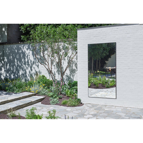 Lucka Outdoor XXL full length garden mirror, 200cm rectangular black metal frame, mounted on a white brick exterior wall reflecting garden greenery and a stone patio.