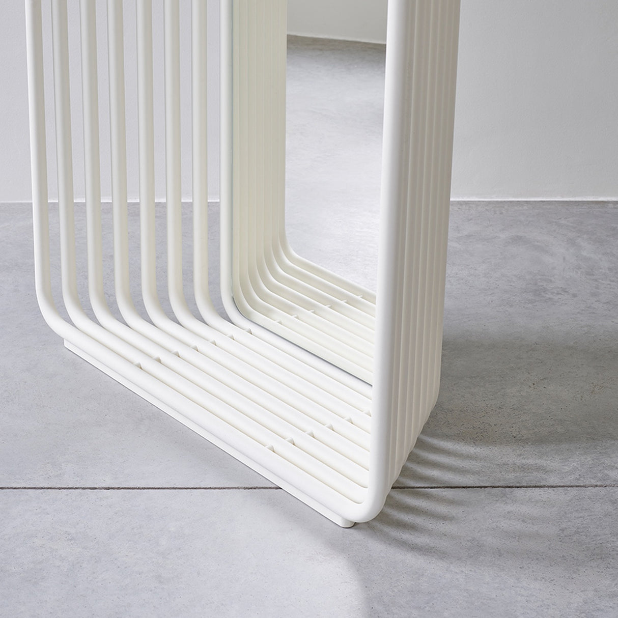 Close-up of the Echo White free standing mirror's base, showing stacked white powder-coated metal slats curving round with a mirrored panel between, set on a concrete floor.