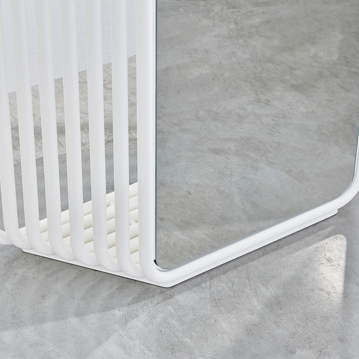 Close-up of the Echo White Free Standing Mirror's base, showing white powder coated metal slats curving round to support the mirrored panel, set on a concrete floor.