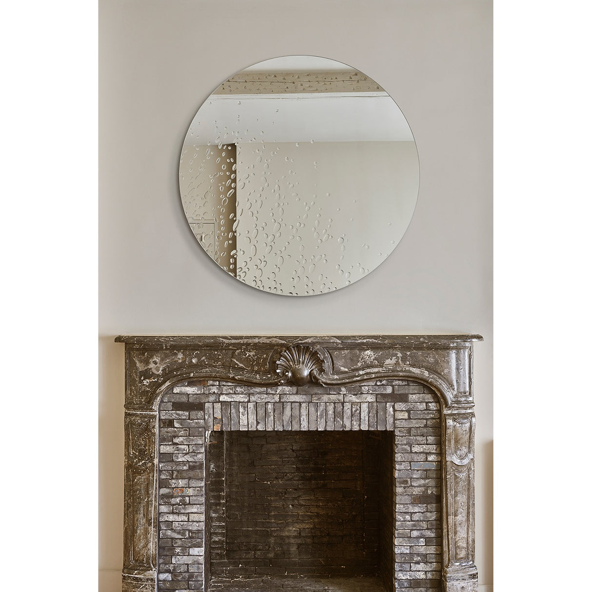 Frameless round wall mirror with hand-applied dewdrop texture on silvered glass, mounted above a stone fireplace with carved shell detail.