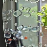 Matin 60cm round wall mirror with hand-applied dewdrop details along the lower edge, reflecting a bright window and greenery.