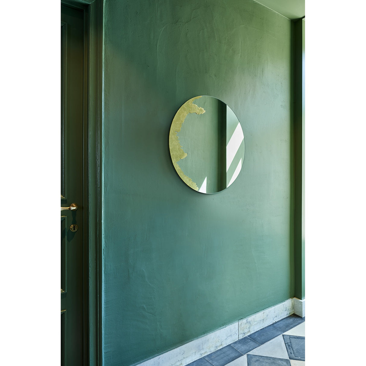 Ornato Gold Leaf Round Rotating Wall Mirror 70cm, frameless round glass with hand-applied gold leaf patch on one edge, mounted on a green wall in a hallway setting.